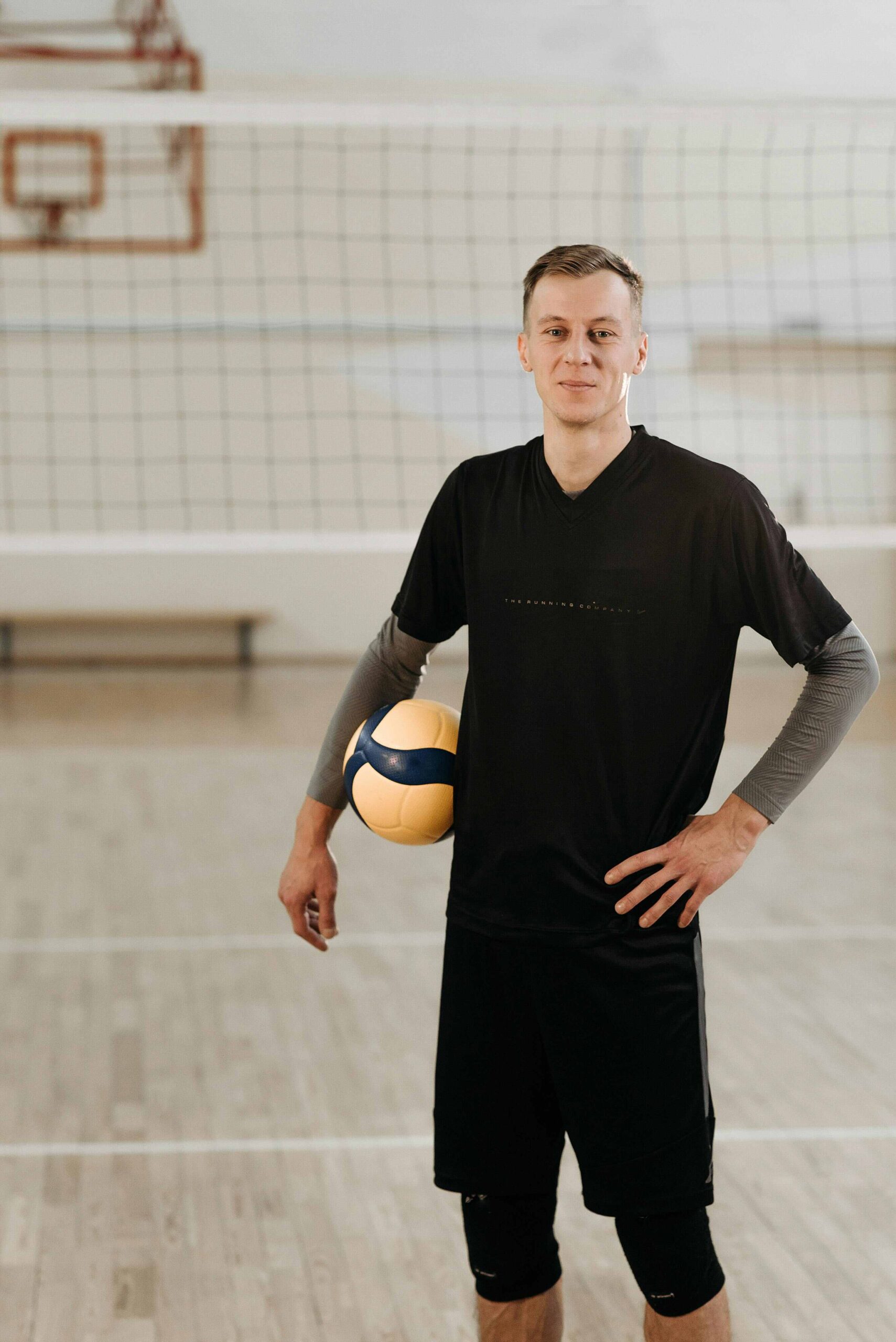 A man in athletic wear stands on an indoor volleyball court, holding a yellow and blue volleyball under one arm and smiling at the camera.