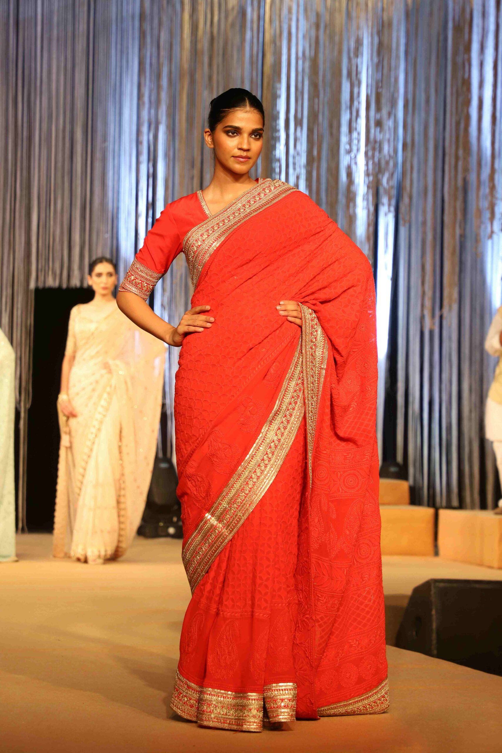 A model confidently walks the runway wearing a vibrant red saree with gold borders, posing with one hand on her hip. Other models in light-colored sarees are visible in the background.
