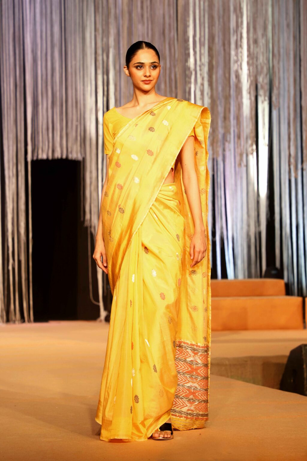 A woman walks a runway wearing a bright yellow saree with subtle silver patterns and a decorative border. The background features vertical silver streamers and soft, warm lighting.