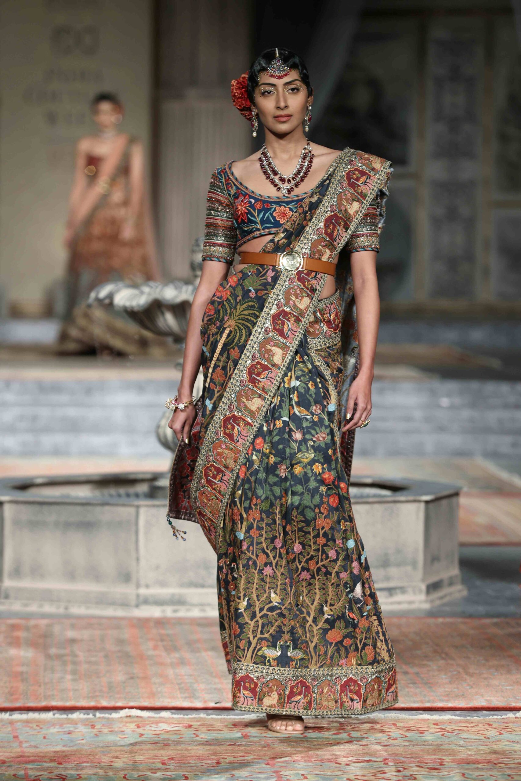 A model walks the runway wearing a richly embroidered, traditional Indian saree with floral and elephant motifs, paired with a matching blouse, jewelry, and a decorative hair accessory. The background shows another model and ornate decor.