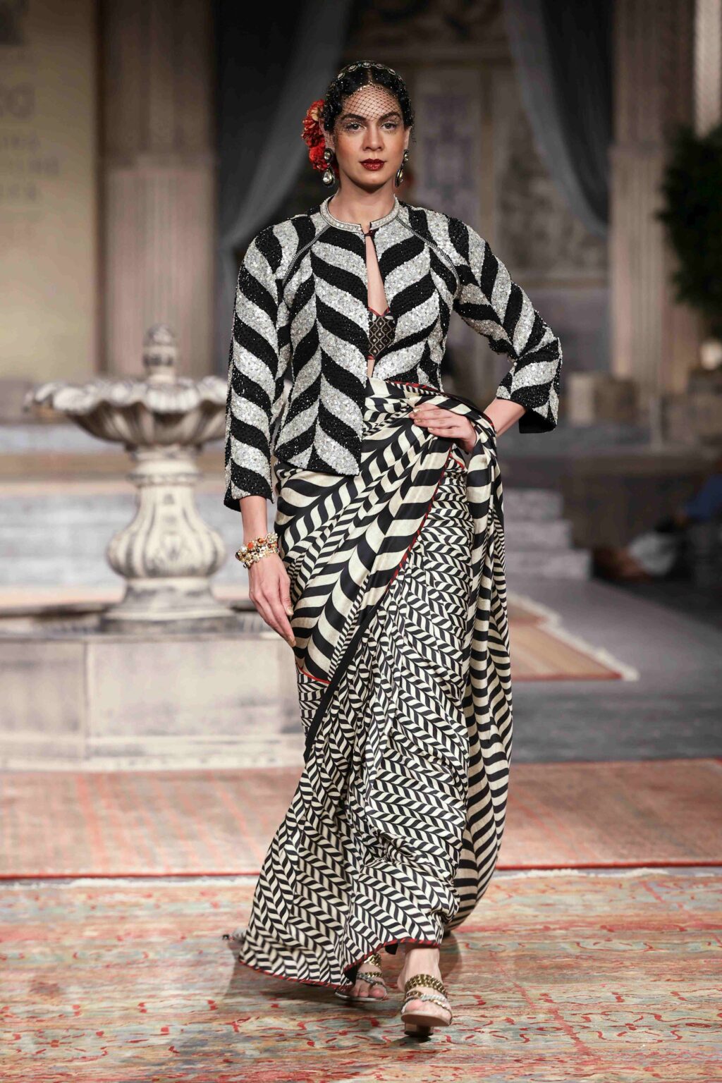 A woman models a black and white patterned saree with a matching jacket, walking on a runway in an ornate setting with a fountain and columns in the background.