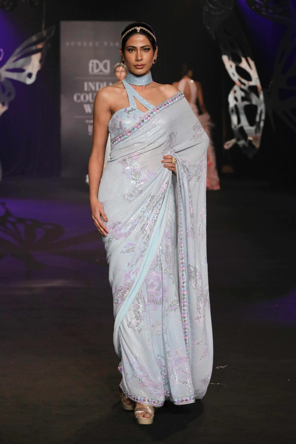 A model walks a runway wearing a light blue, floral-patterned saree with a matching blouse, accessorized with a headband and earrings, against a dark background with butterfly decorations.