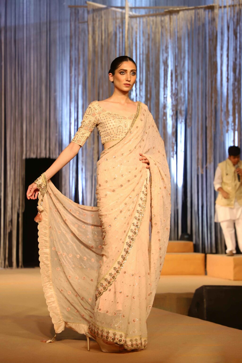 A model walks on a runway wearing an elegant, light-colored saree with intricate embroidery, while holding one end of the saree. The background features vertical metallic streamers and a man standing in traditional attire.