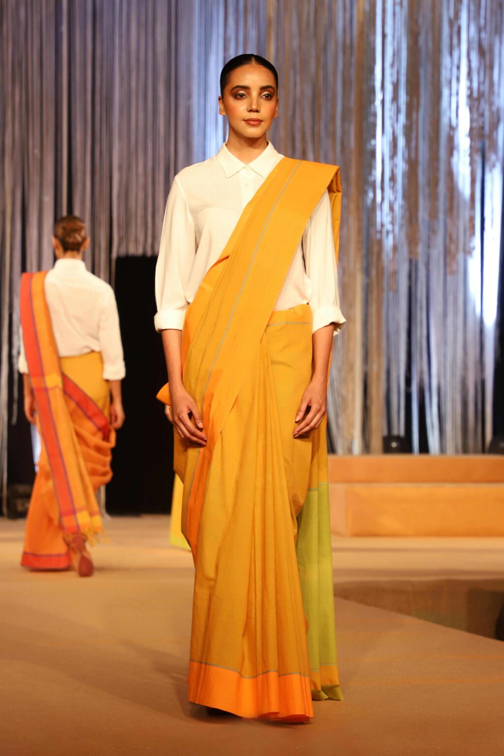 A model walks a runway wearing a mustard yellow, orange, and green sari paired with a white blouse. Another model in a similar outfit is visible in the background. The setting has vertical silver drapes.