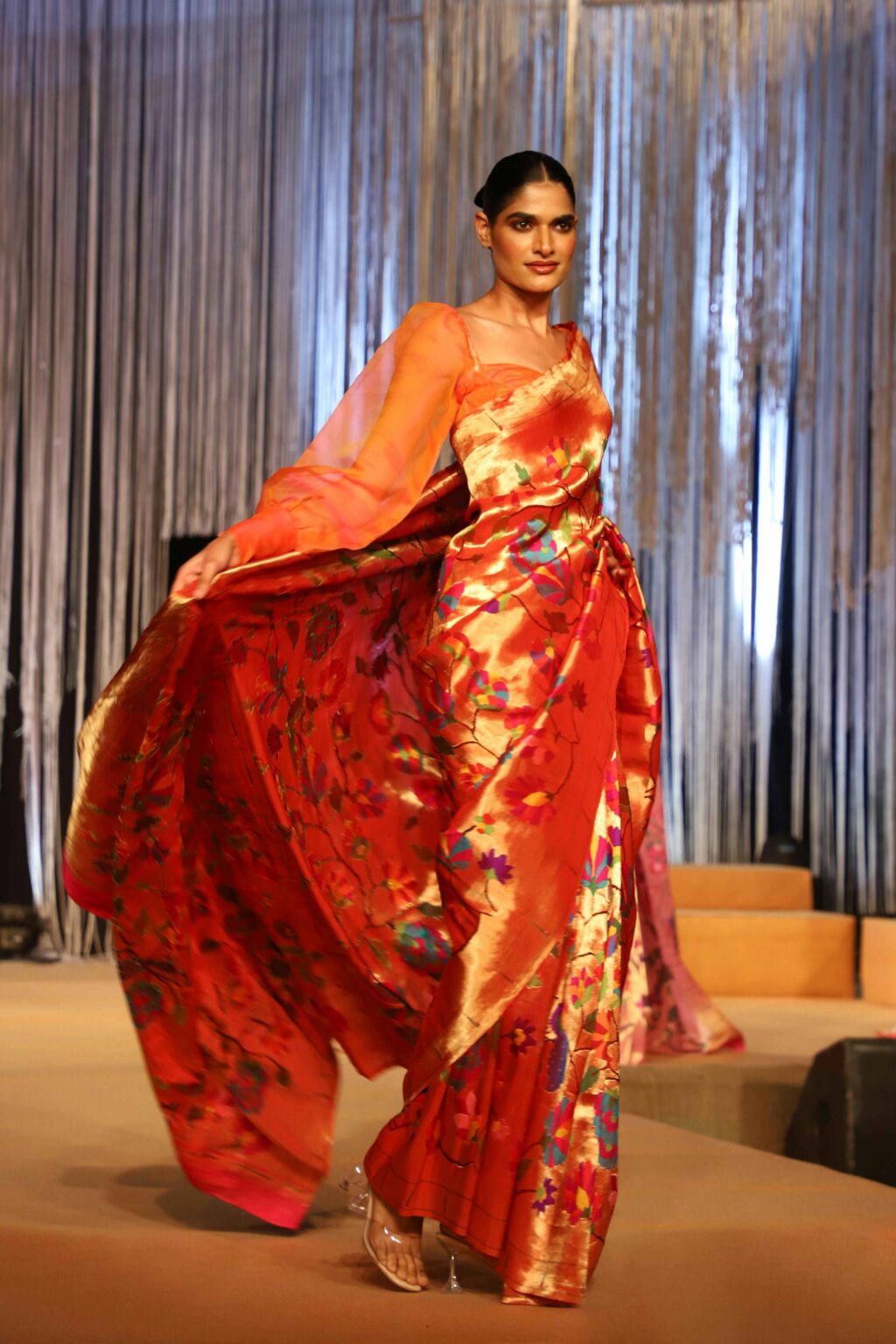 A model walks a runway wearing a vibrant orange and gold saree with floral patterns, flowing sheer sleeves, and gold heels. The background features vertical silver streamers and a warm-lit stage.