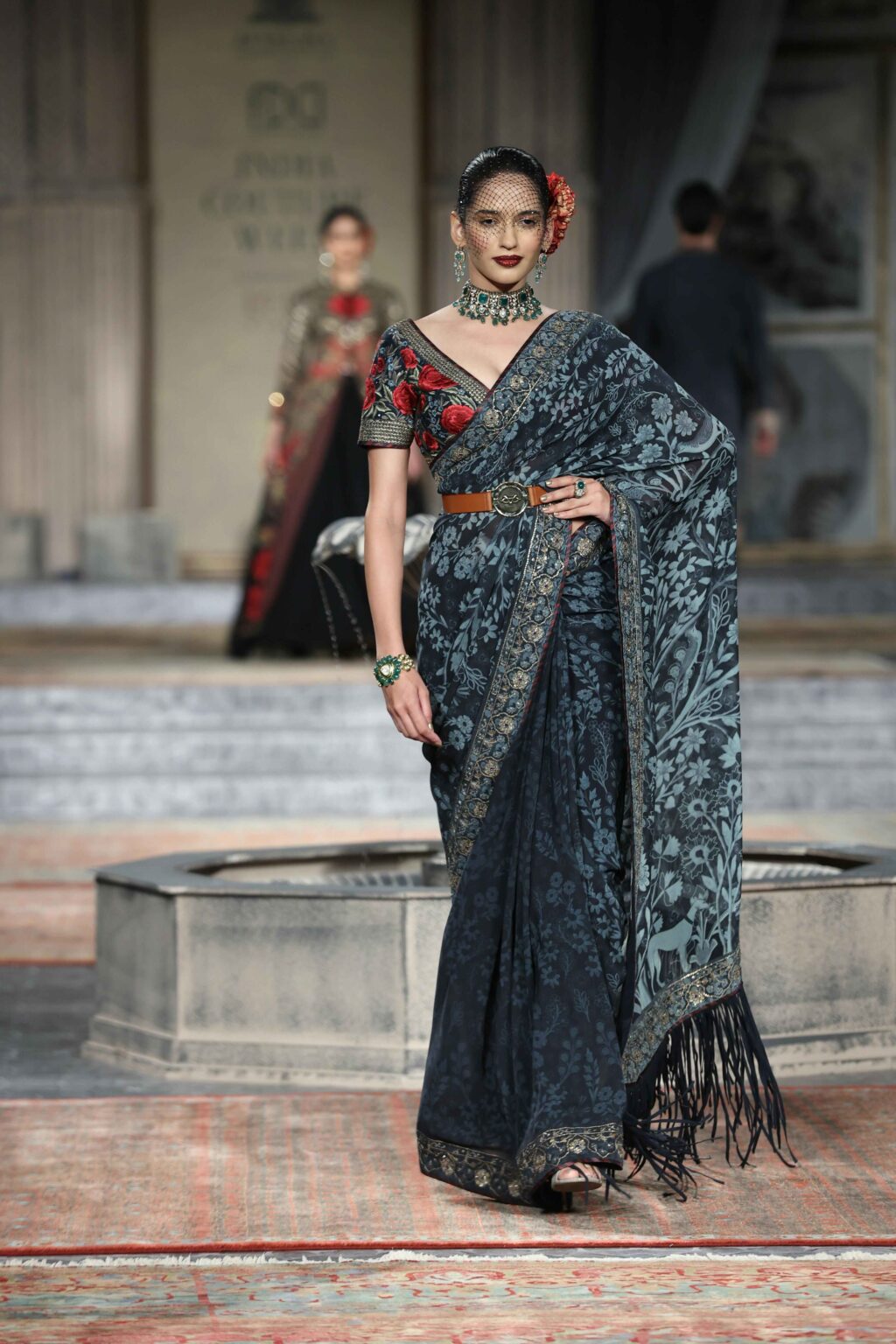 A model walks a runway wearing a dark blue, floral-patterned saree with a black fringe, a brown belt, and a red floral blouse. She accessorizes with jewelry, a hair flower, and a netted veil. Other models appear in the background.