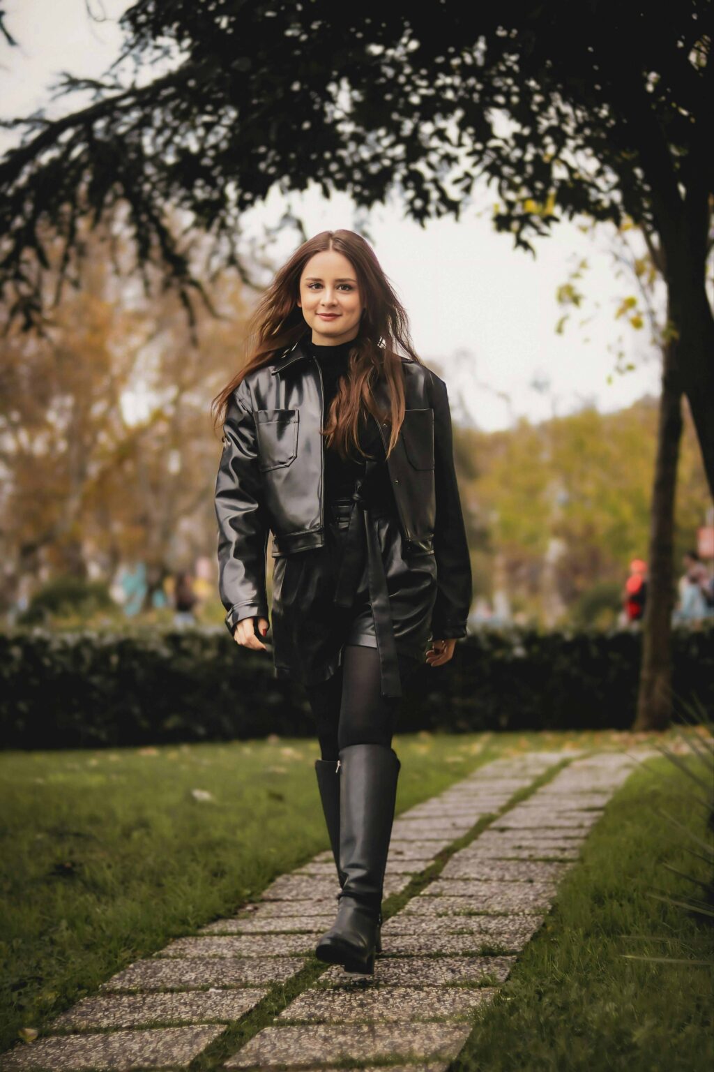 A woman with long brown hair walks confidently on a stone path in a park, wearing a black leather jacket, black skirt, and black boots. Trees and greenery surround the walkway.
