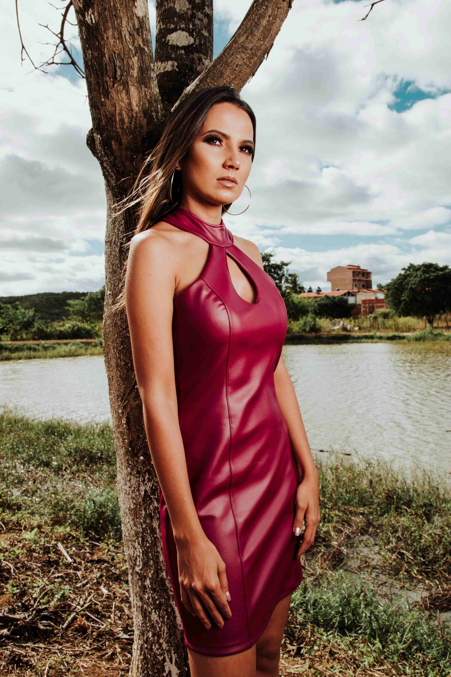 A woman in a sleeveless maroon dress stands in front of a tree by a lake, with greenery and cloudy sky in the background.