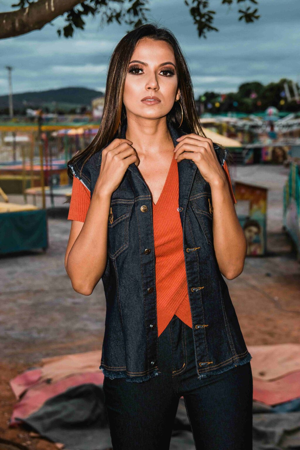 A woman with long dark hair wears a denim vest over an orange top, standing outdoors with colorful carnival rides blurred in the background. She looks directly at the camera, holding her vest with both hands.