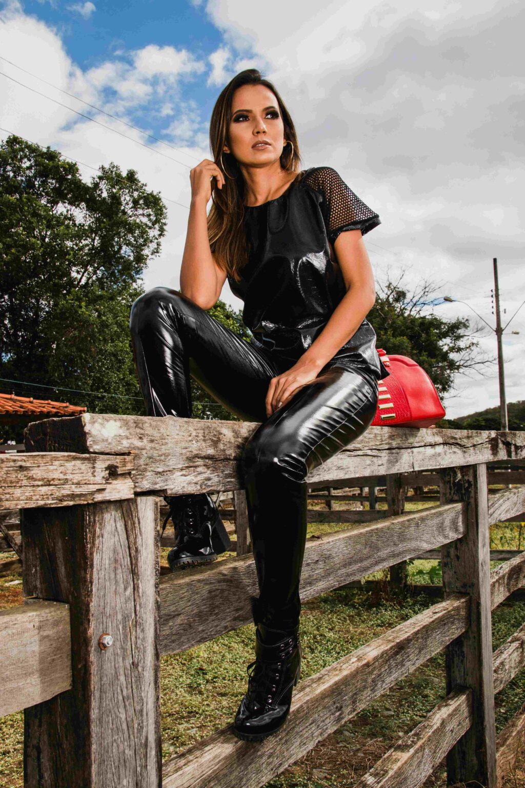 A woman in shiny black pants and a black mesh top sits on a rustic wooden fence outdoors, with trees and a cloudy sky in the background and a red bag beside her.