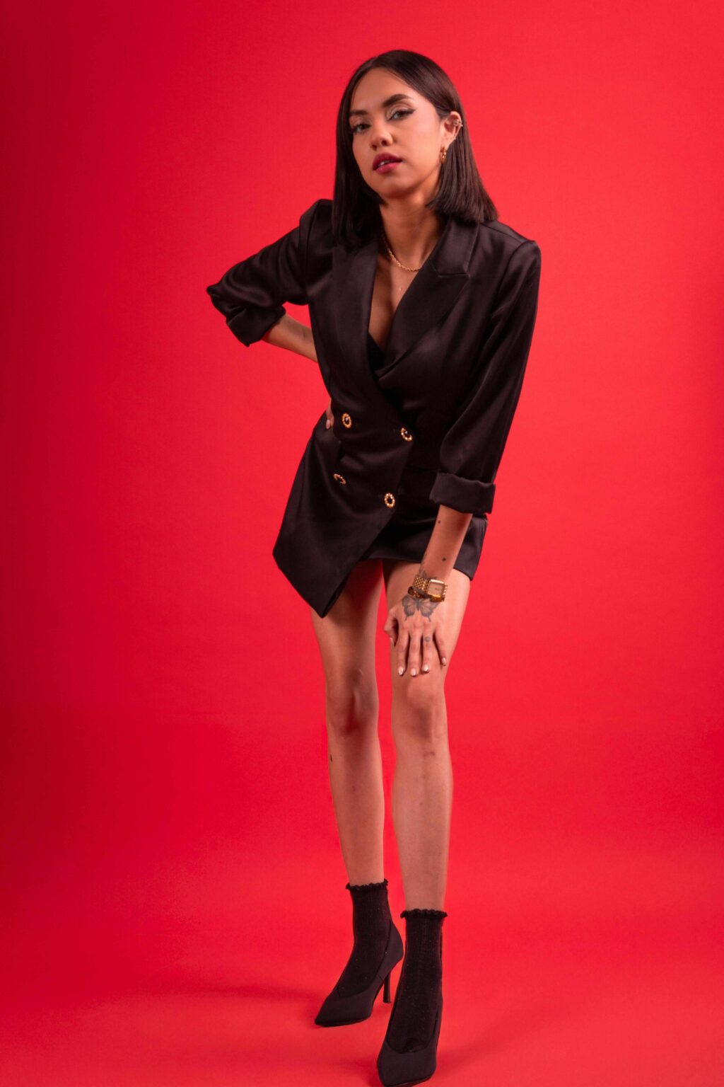 A woman poses against a red background, wearing a black blazer dress with gold buttons, black ankle boots, and statement rings, looking confidently at the camera with one hand on her knee.