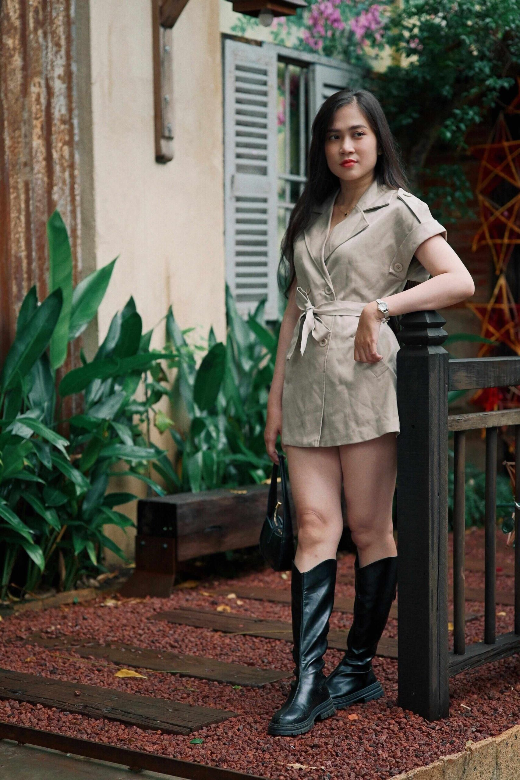 A woman with long dark hair, wearing a beige belted romper and black knee-high boots, stands on a wooden bridge. She holds a handbag and leans on a black railing. Green plants and a rustic building are in the background.