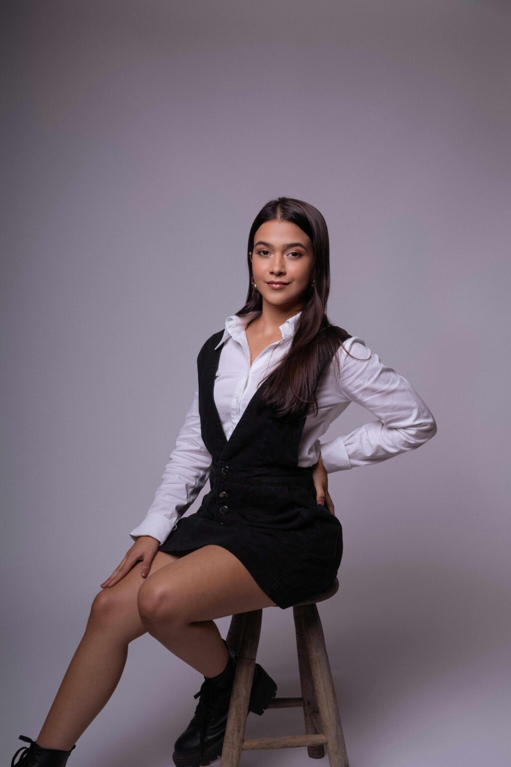 A young woman with long dark hair wearing a white blouse and black dress sits confidently on a wooden stool against a plain, light background.