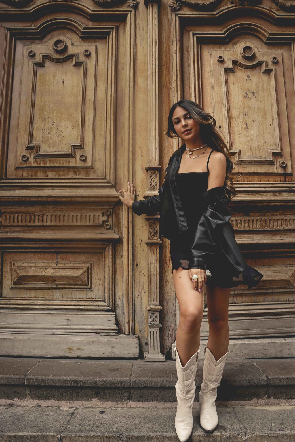 A woman wearing a black outfit, black jacket, and white cowboy boots stands confidently in front of large, ornate wooden doors, resting one hand on the door and looking at the camera.