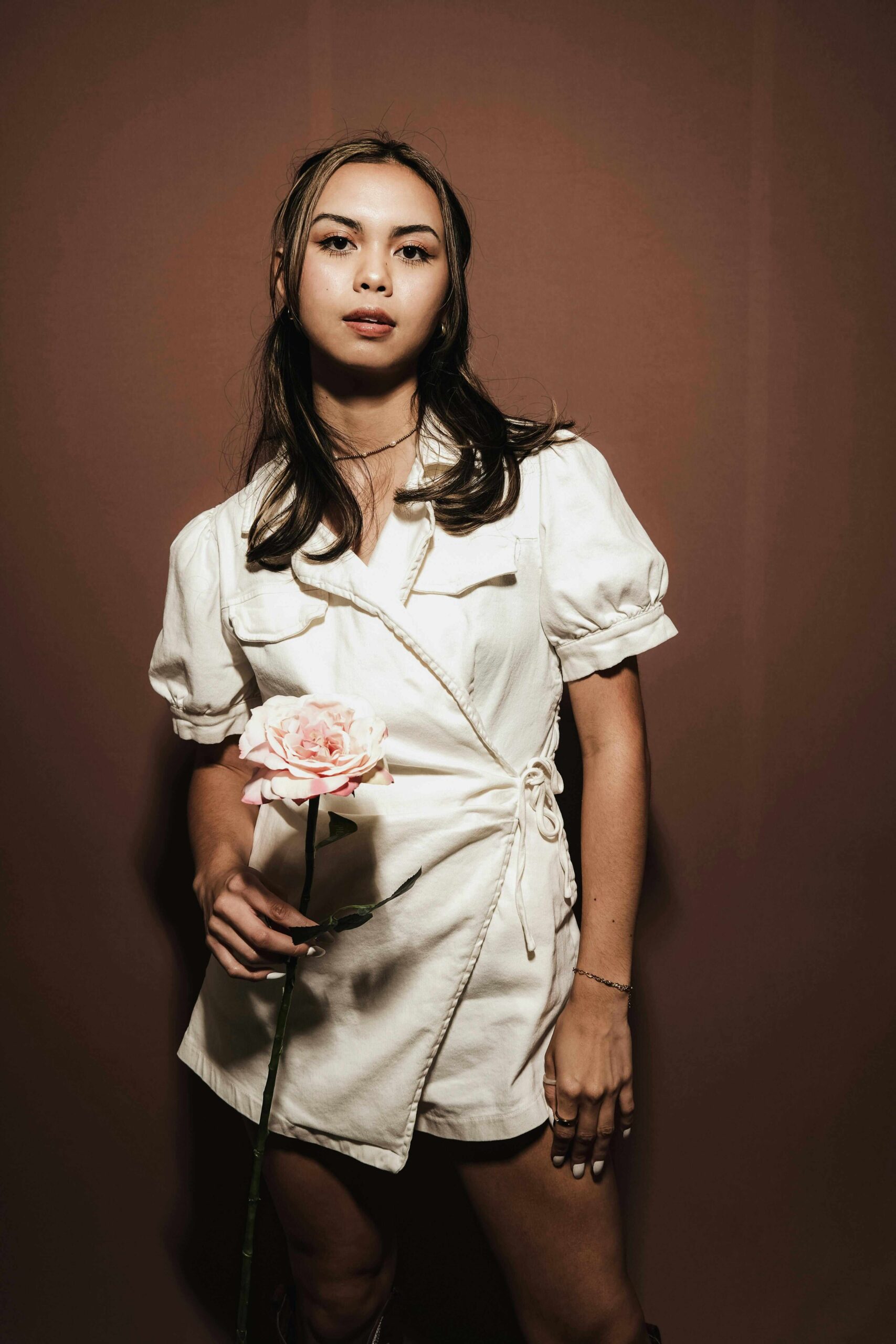 A woman wearing a short-sleeved, white wrap dress stands against a brown background, holding a pink rose in one hand and looking confidently at the camera.